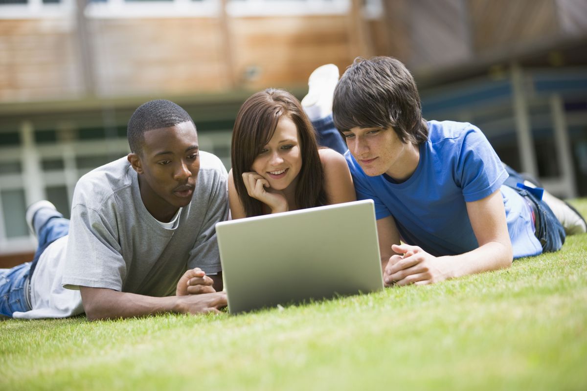 zur Vergrößerungsansicht des Bildes: Drei junge Erwachsene liegen vor einem Laptop auf dem Rasen.