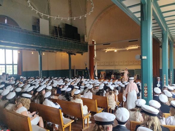 Es sind Abiturient*innen in einer großen Halle von hinten zu sehen. Sie sitzen auf Holzbänken in Reihen und Tragen weiße Roben und weiße Hüte.