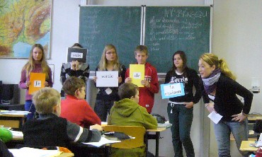 zur Vergrößerungsansicht des Bildes: Das Bild zeigt eine Unterrichtssituation mit einer Praktikantin sowie Schülerinnen und Schülern.