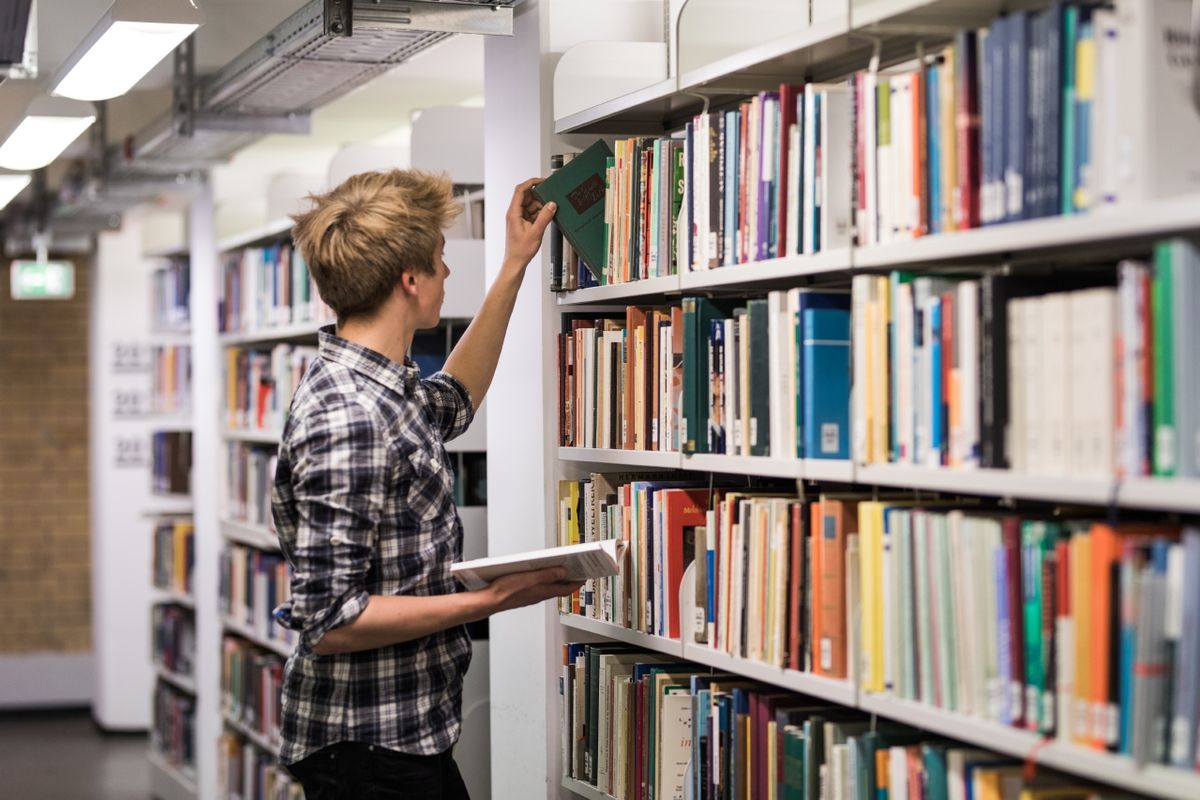 zur Vergrößerungsansicht des Bildes: Ein Student steht in einem Gang und greift in ein Bibliotheksregal nach einem Buch während ein anderes Buch in der Hand hält, Foto: Christian Hüller