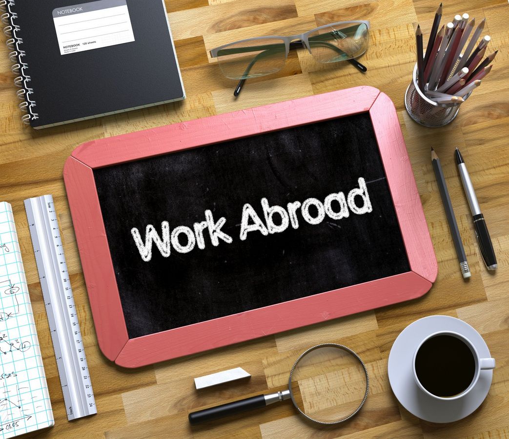 Auf einem Schreibtisch liegt neben Stiften und anderen Dingen eine Tafel mit den Worten "Work Abroad".
