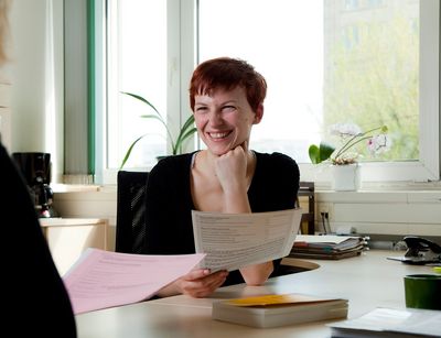 Foto: zwei Frauen sitzen sich an einem Schreibtisch in einem Beratungsgespräch gegenüber und haben Zettel in der Hand