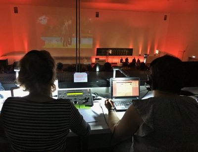 Blick aus dem Regieraum in den rötlich beleuchteten Vortragssaal. Zwei Techniker sitzen vor Computer und Mischpult.