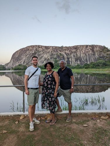 Auf dem Bild ist Lukas Fiedler in Begleitung eines mosambikanischen Paares beim Besuch der Montes Nairuco in Namula zu sehen. Sie stehen am Geländer vor einer Wasserfläche, hinter der sich der Berg Nairuco erhebt. Im Wasser ist eine Spiegelung des Berges zu sehen.