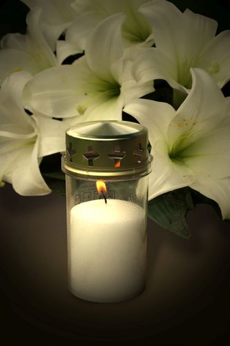 Es handelt sich hierbei um ein Stillleben mit weißer Grabkerze und weißen Lilien auf dunklem Untergrund. Die Kerze ist angezündet und steht vor den liegenden Blumen.