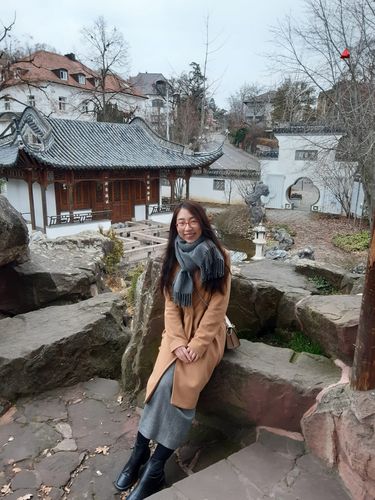 Jie Li an einem Stein in einem japanischen Garten lehnend. Sie ist in Schal und dicker Jacke gekleidet, der Himmel ist bewölkt, es scheint Winter zu sein. Im Hintergrund sieht mein ein japanisches Holzhaus und mehr Bäume ohne Blätter, Steine und einen kleinen Teich.