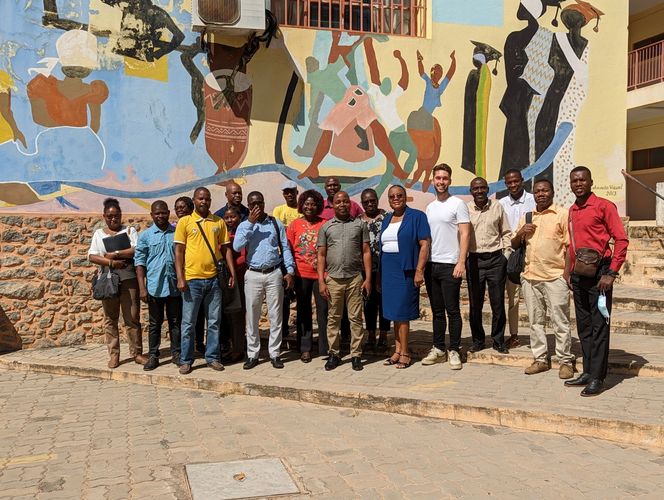 Gruppenfoto von Lukas Fiedler mit einigen Angehörigen der Universidade de Nampula vor einer bunt bemalten Wand. Auf der Wand sind afrikanische Motive zu sehen.