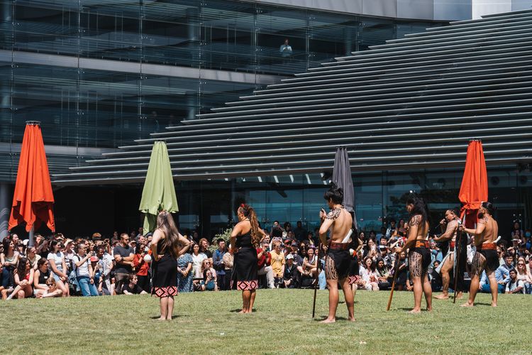 Abbildung einer Veranstaltung von Maori im Vordergrund während im Hintergrund ein Teil des Universitätsgebäudes abgebildet ist.