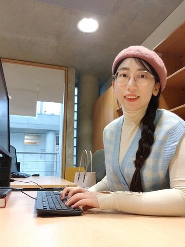 Zu sehen ist Jie Li an ihrem Arbeitsplatz. Sie schaut zur Seite in die Kamera, während sie auf der Laptop Tastatur tippt