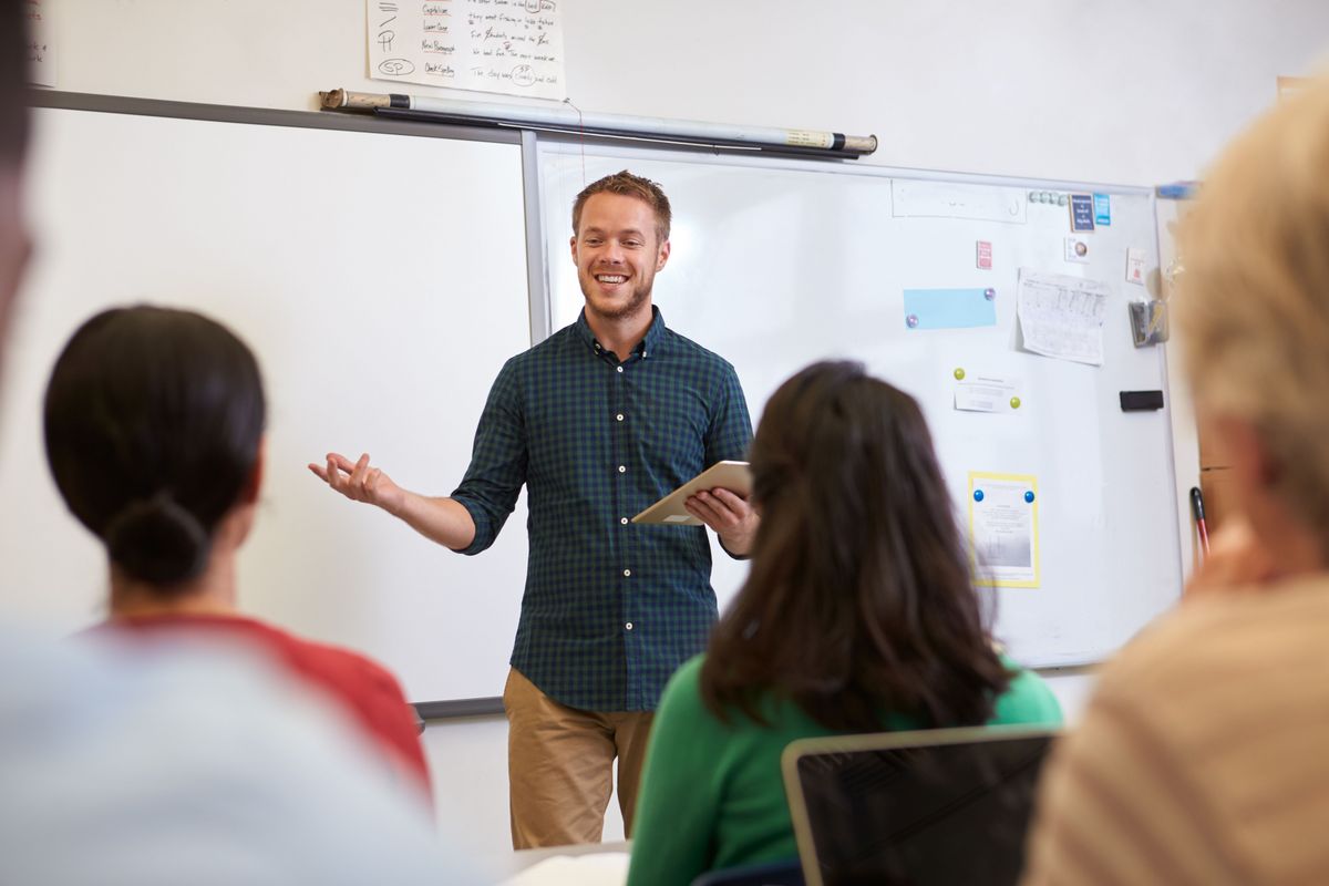 Ein junger Lehrer steht lächelnd vor einer Klasse.