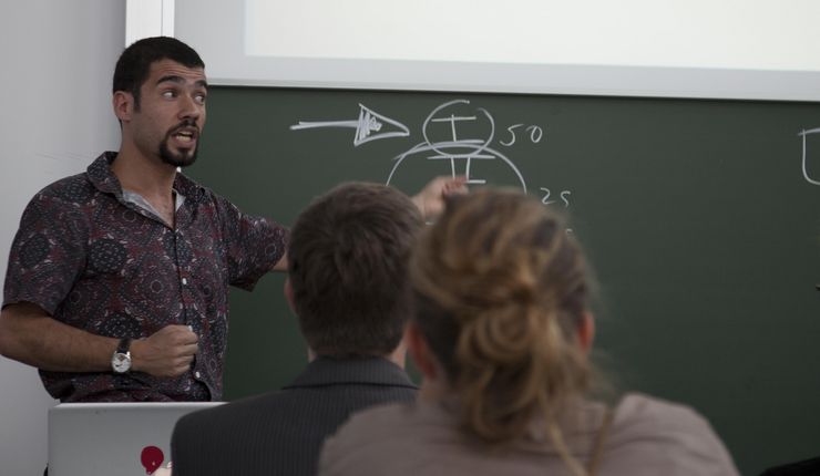 A lecturer stands in front of a blackboard with writing on it and explains something to the students sitting in front of him.