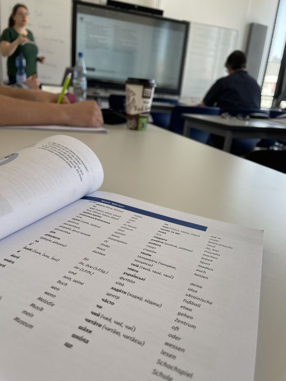 Ein aufgeschlagenes Wörterbuch aus dem Kurs liegt auf dem Tisch. im Hintergrund verschwommen ist der Kurs zu erkennen.