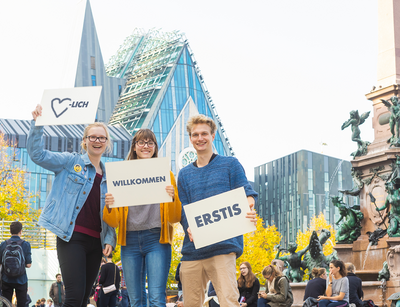 3 Studierende stehen auf dem Leipziger Augustusplatz und halten Schilder mit dem Aufdruck "Herzlich Willkommen Erstis" hoch.