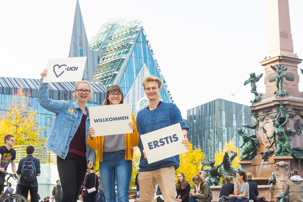 Auf dem Bild sind 3 junge Menschen auf dem Leipziger Augustusplatz zu sehen. Sie halten Schilder mit dem Aufdruck "Herzlich Willkommen Erstis" hoch.
