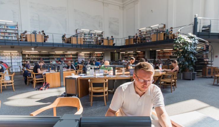 Studierende in der Bibliothek, im Vordergrund ein Student am Schreibtisch, im Hintergrund sind weitere Arbeitsplätze zu sehen
