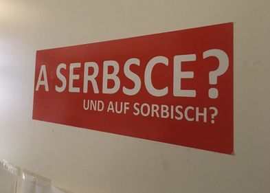Auf dem Bild ist ein Schild zu sehen, auf dem "Und auf Sorbisch? auf Deutsch und Sorbisch steht