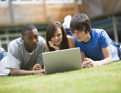 Drei junge Erwachsene liegen vor einem Laptop auf dem Rasen.