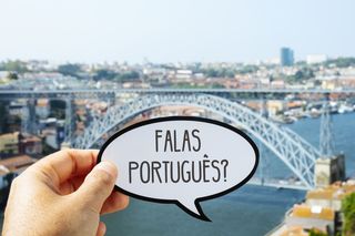 Auf dem Bild ist eine verschwommene Silhouette einer Stadt zu sehen, die an einem Fluss liegt, der von einer Stahlgerüstbrücke überspannt wird. Im Vordergrund ist eine Hand zu sehen, die ein Schild in Form einer Sprechblase hält und auf dem steht: Falas português?