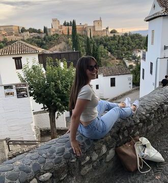 Man sieht Lilia auf einer Steinmauer sitzen, während im Hintergrund landestypische Häuser und kleine Gassen, so wie die Alhambra zu sehen sind. Lilia trägt ein kurzes Shirt und eine Sonnenbrille.