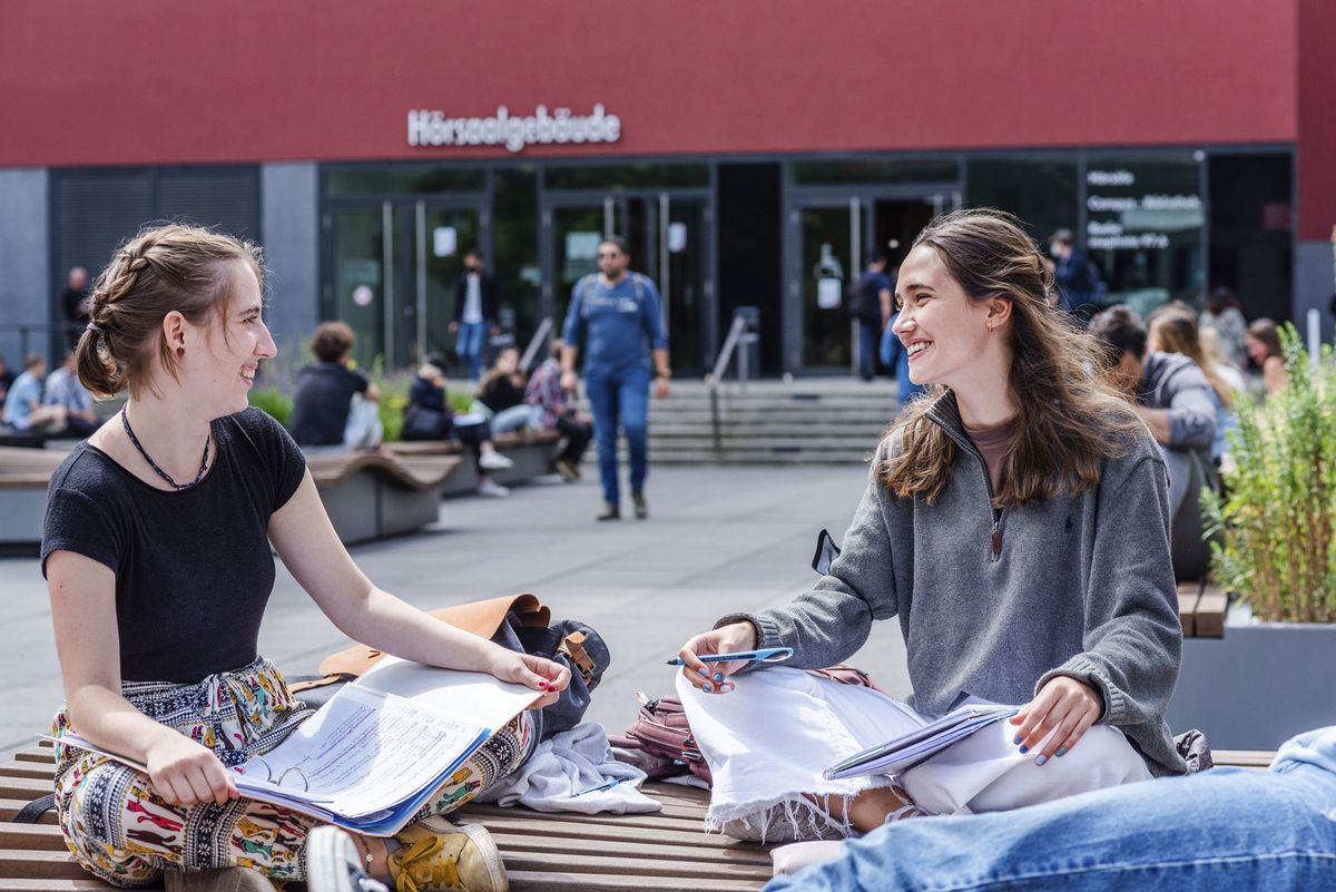 Two female students sit cross-legged on a bench in the university courtyard and talk. In the background, people walking into the lecture hall building can be seen out of focus.